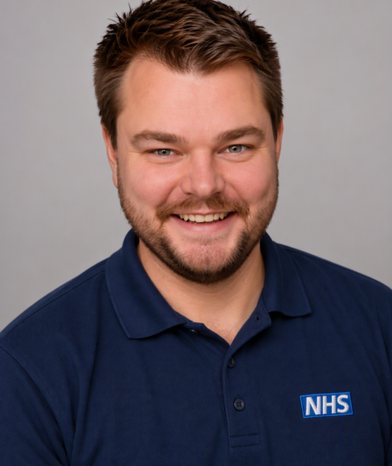 A headshot of a smiling man with short brown hair, a light beard, and a mustache, wearing a dark blue polo shirt featuring an NHS logo on the chest.