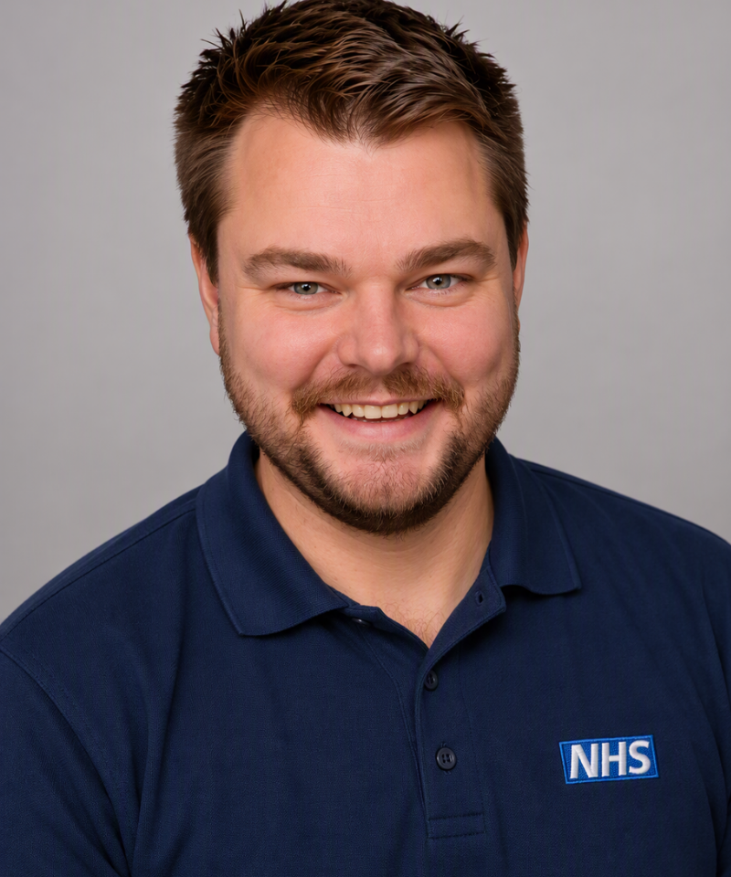 A headshot of a smiling man with short brown hair, a light beard, and a mustache, wearing a dark blue polo shirt featuring an NHS logo on the chest.