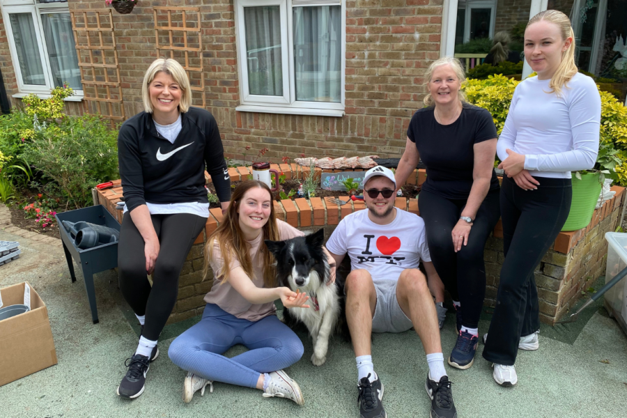 Five people and a border collie dog pose outdoors in front of a brick building garden bed. Two women are seated on the left wall: one wearing a black Nike sweatshirt and black leggings, and another sitting cross-legged on the ground below her feeding the dog. Next to them on the ground, a young man wearing a white "I ❤️ NY" t-shirt, white cap, and sunglasses sits beside the dog. Sitting on the raised brick edge behind him is a blonde woman in a black t-shirt, and standing to her right is a young woman wearing a long-sleeved white top and dark pants.