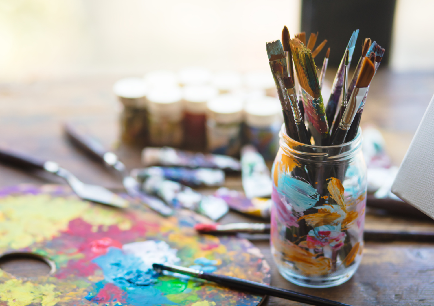 Selection of paintbrushes in a glass jar alongside a colorful paint palette and art supplies on a wooden table.