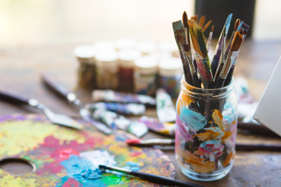 Selection of paintbrushes in a glass jar alongside a colorful paint palette and art supplies on a wooden table.