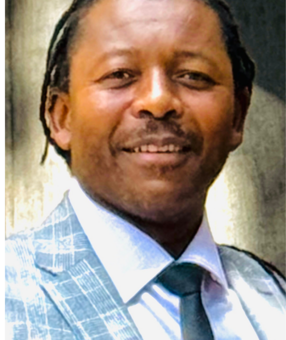 Headshot of George Garamukanwa wearing a light grey plaid suit jacket, white shirt, and black tie.
