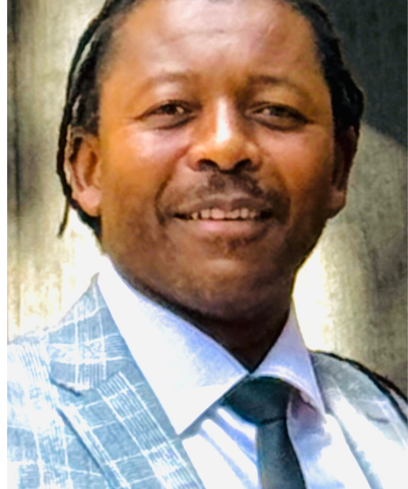 Headshot of George Garamukanwa wearing a light grey plaid suit jacket, white shirt, and black tie.