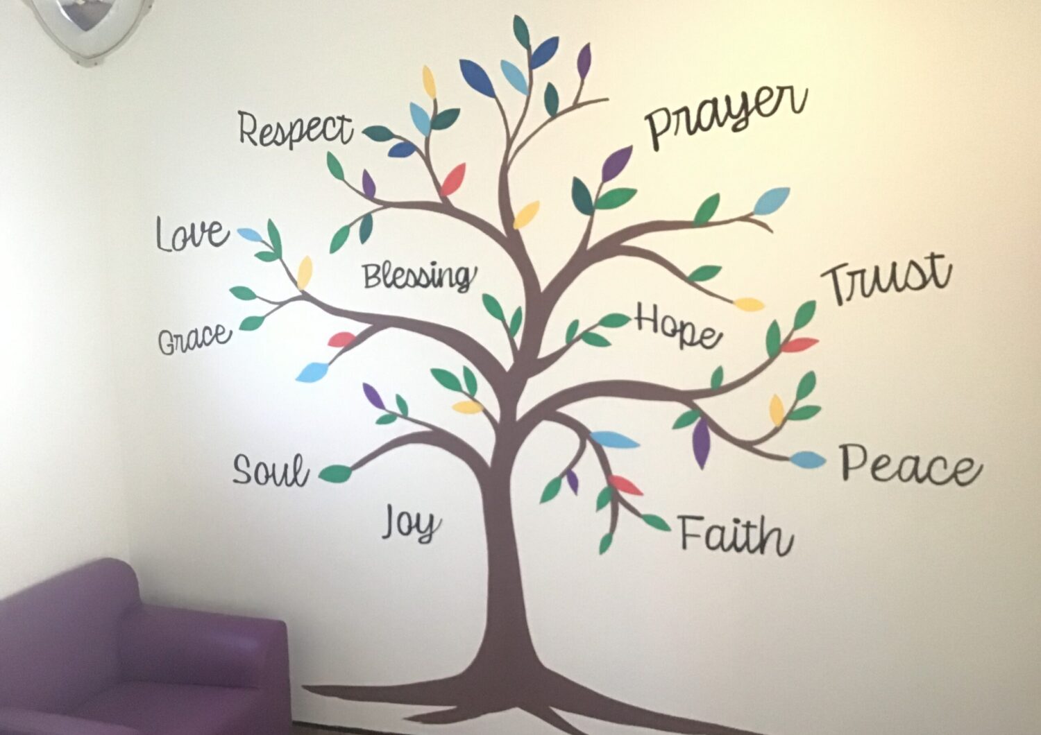 A wall decal featuring a stylized brown tree with multi-colored leaves painted on a light cream wall. Cursive text featuring uplifting words surrounds and intertwines with the branches, including 