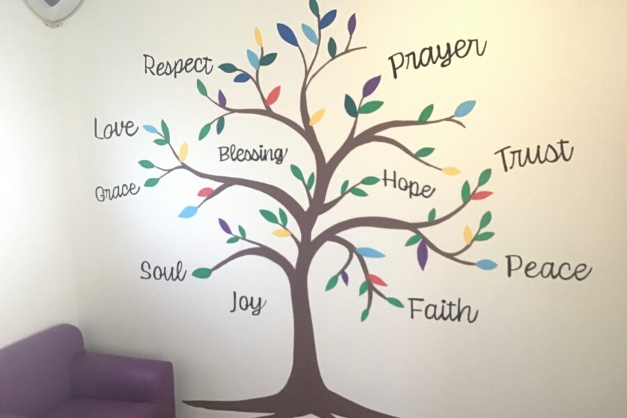 A wall decal featuring a stylized brown tree with multi-colored leaves painted on a light cream wall. Cursive text featuring uplifting words surrounds and intertwines with the branches, including "Respect," "Love," "Grace," "Soul," "Blessing," "Joy," "Prayer," "Hope," "Trust," "Faith," and "Peace". A light fixture is mounted on the wall to the top left, and part of a purple armchair is visible in the bottom-left corner of the room.