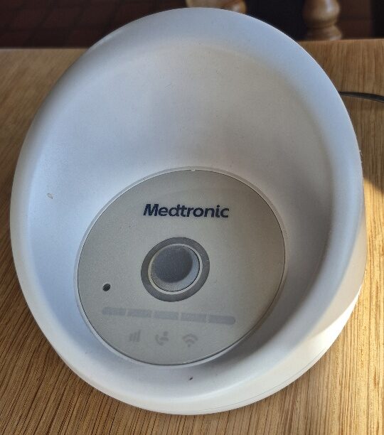A close-up view of a white, bowl-shaped Medtronic CareLink monitor sitting on a light wooden table surface. The device features a circular center faceplate displaying the Medtronic logo, a central power button, and indicator icons for cellular signal, status, and Wi-Fi connection.