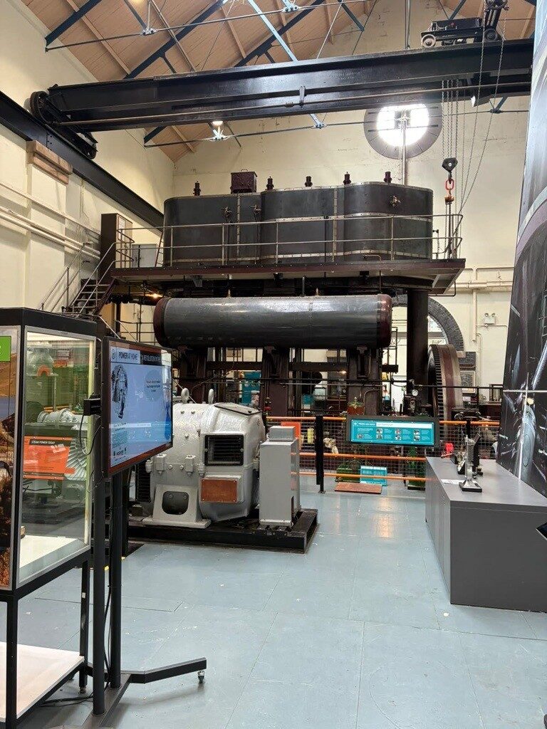 An interior view of a industrial museum hall, featuring large restored power machinery. In the center stands a massive dark metal steam engine structure with an upper walkway platform and a horizontal cylindrical tank below it. In the foreground, there is a grey industrial generator casing alongside interactive digital information screens and display cases detailing industrial power history. Overhead, an industrial crane beam spans the vaulted hall ceiling.