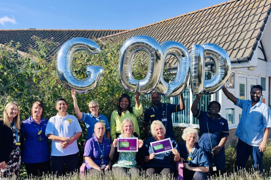 A group of staff members standing and kneeling outdoors in front of a building, celebrating a "Good" CQC rating. Large metallic foil balloons spelling out "GOOD" are held aloft across the back row.