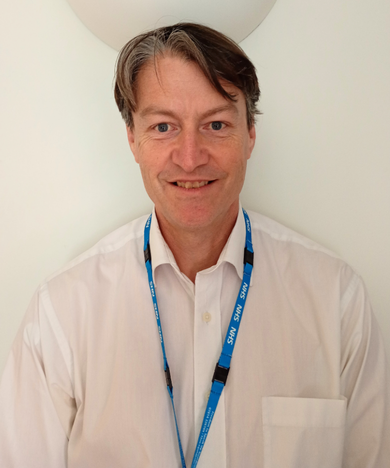 A portrait of a man, identified as Simon Morgan, facing forward and smiling slightly against a plain white background. He has short, dark brown hair with graying sides parted to the left, blue eyes, and light skin. He is wearing a white button-down shirt with a breast pocket and a bright blue lanyard around his neck featuring white lettering and black clips.