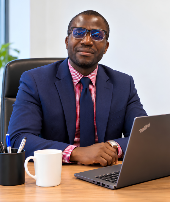 A portrait of a man, identified as Sunday Adebayo, seated at a wooden desk and looking directly at the camera with a gentle smile. He is wearing a dark blue suit jacket, a red and white checkered dress shirt, a solid blue tie, and dark-rimmed glasses. On the desk in front of him are a open grey ThinkPad laptop, a white ceramic mug, and a black pen holder with several pens.