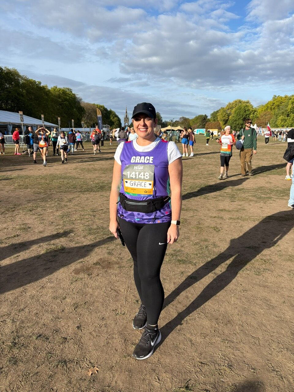 A woman stands outdoors in a grassy park, smiling at the camera, during what appears to be a running event or race. She is wearing a purple running top over a white short-sleeved shirt, black leggings, a black cap, and running shoes, with a race bib pinned to her chest displaying the name 