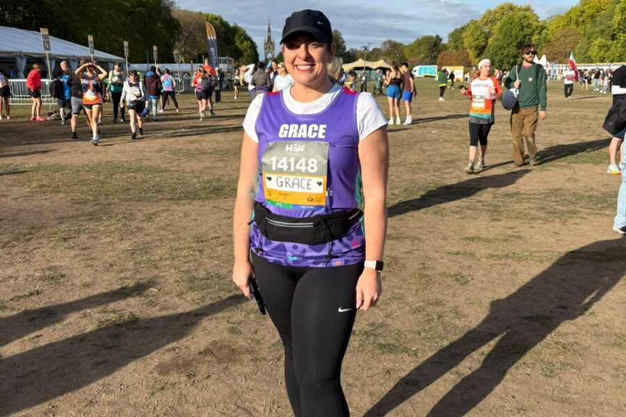A woman stands outdoors in a grassy park, smiling at the camera, during what appears to be a running event or race. She is wearing a purple running top over a white short-sleeved shirt, black leggings, a black cap, and running shoes, with a race bib pinned to her chest displaying the name "GRACE" and number "14148". In the background, other people are scattered across the grassy field under a partly cloudy blue sky, with trees and tents visible in the distance. Long shadows stretch across the ground in the afternoon light.
