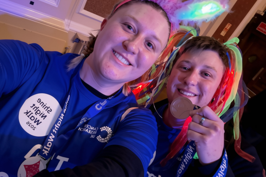 Two smiling women take a selfie wearing matching blue Cancer Research UK "Shine Night Walk 2024" t-shirts and lanyards. The person on the right holds up a finisher's medal and wears colorful light-up headgear, while the person on the left wears light-up bunny ears under indoor lighting.