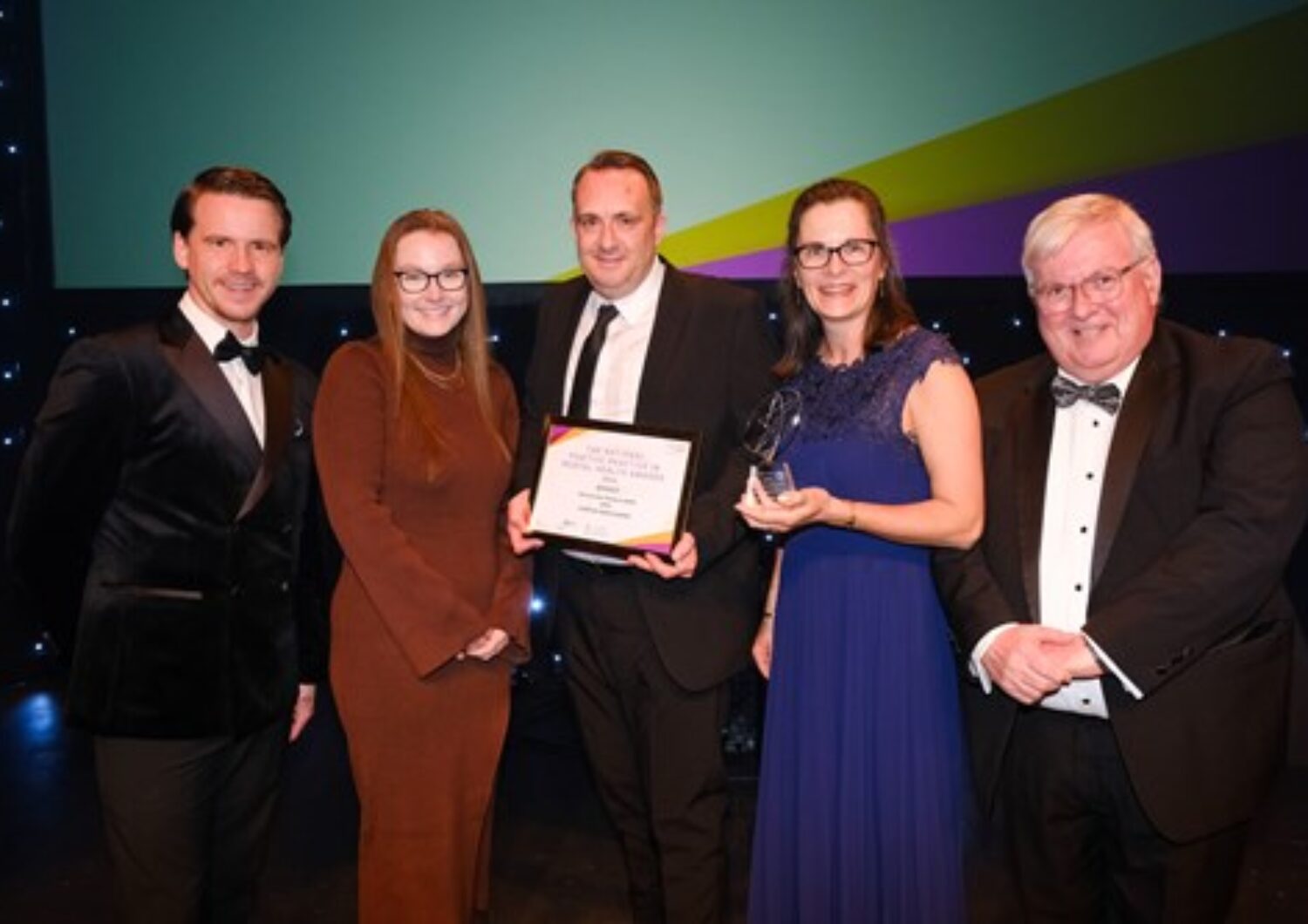 Three men and Two women smiling at a camera holding their team's award.