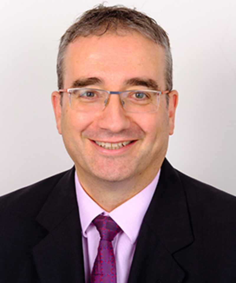 Photo of Nigel Leonard wearing a black suit jacket, pink shirt and tie