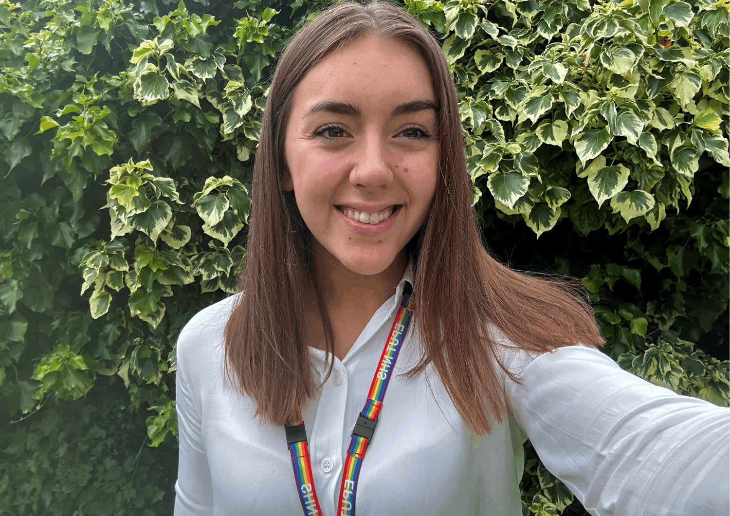 Photo of the volunteer Chloe. She has borwn shulder length hair, is wearing a white shirt and a rainbow lanyard.
