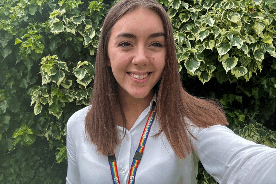 Photo of the volunteer Chloe. She has borwn shulder length hair, is wearing a white shirt and a rainbow lanyard.