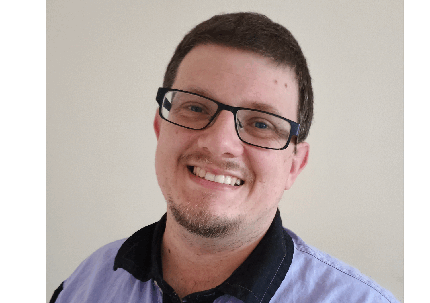 Photo of Dominic smiling at the camera. He has dark brown hair, wearing black rimmed glasses and is wearing a blue shirt.