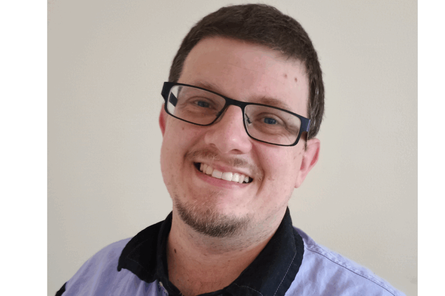 Photo of Dominic smiling at the camera. He has dark brown hair, wearing black rimmed glasses and is wearing a blue shirt.