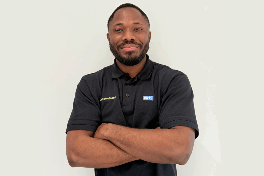 Photo of Harmony smiling at the camera. He is wearing a black t-shirt with the NHS logo on it.