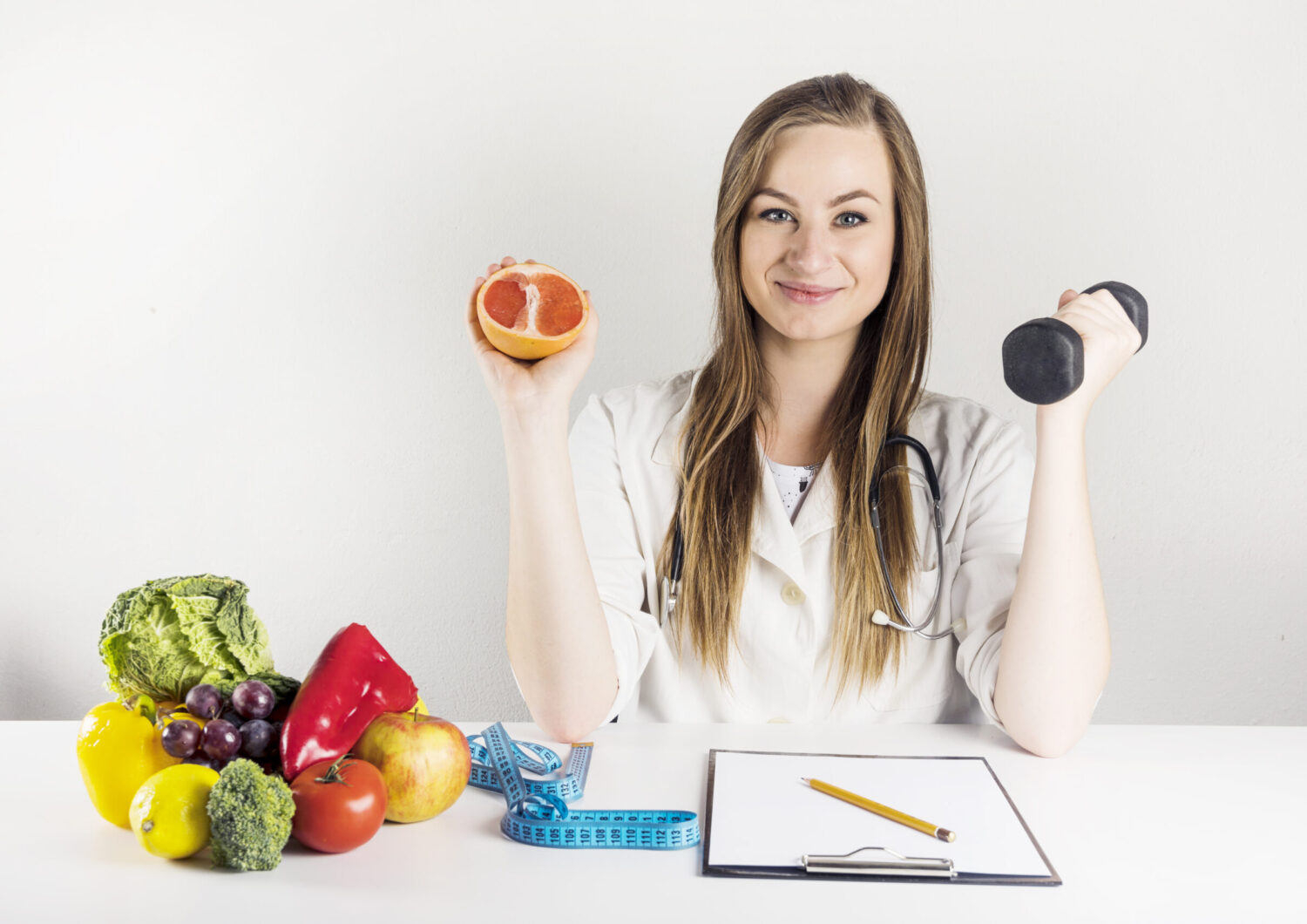 Woman holding half an orange in one hand and a dumbbell in the other. She has a clipboard, fruit and a measuring tape in front of her.