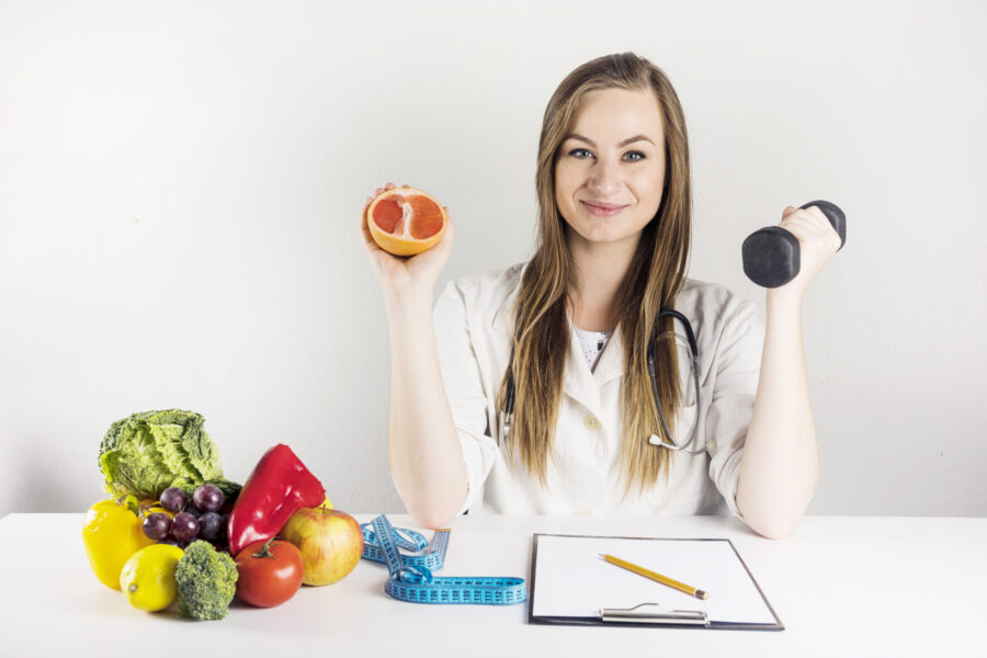Woman holding half an orange in one hand and a dumbbell in the other. She has a clipboard, fruit and a measuring tape in front of her.